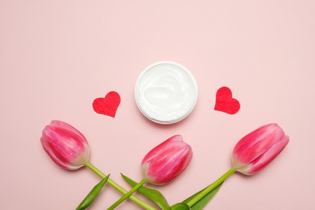 A composition with tulips and a jar of hand cream on a pink background.