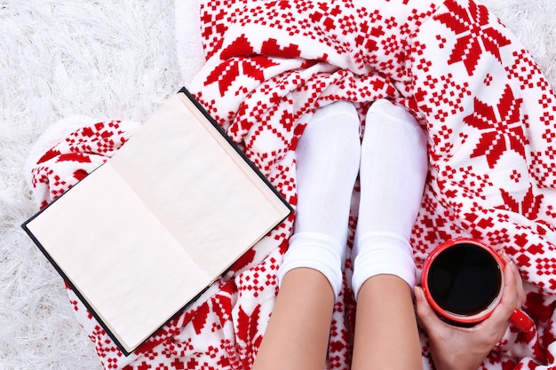 Composition with warm plaid, book, cup of hot drink and female legs, on color carpet background