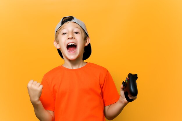 A computer gamer boy on a yellow background shows emotions. a child in a cap and an orange t-shirt is playing a game station