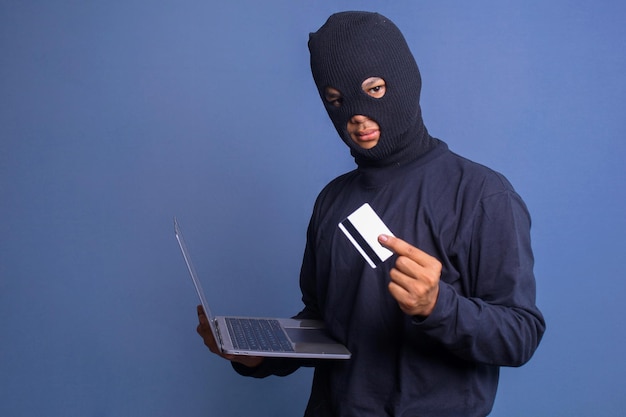 Computer hacker with balaclava holding credit card and stealing data from a laptop Concpet of cyber