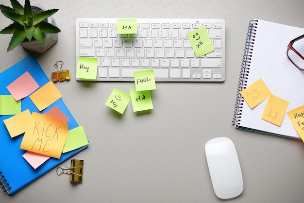 Computer keyboard with stationery and sticky notes on grey. April Fool's Day prank