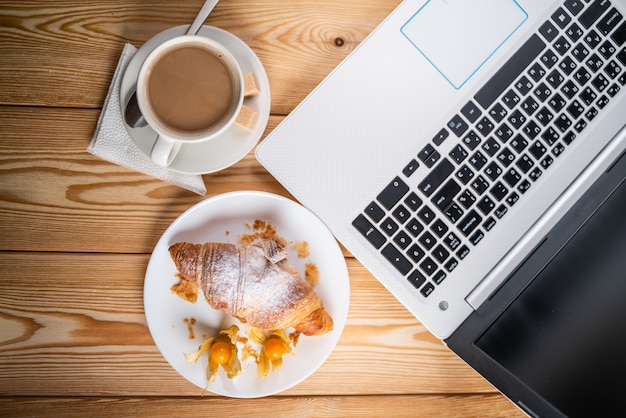 Computer, kop koffie en croissant op bruin houten tafel