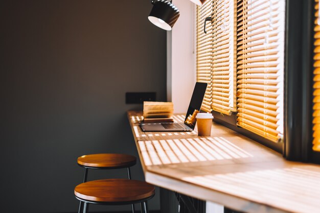 Computer laptop met koffiekopje en open boek over de moderne houten tafel in de keuken in de buurt van raam met gordijn