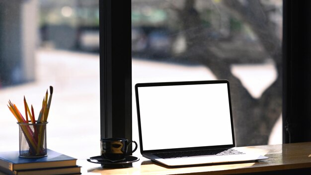 Computer laptop with empty screen, coffee cup, stationery and books on wooden table. Blank screen for your advertising text.