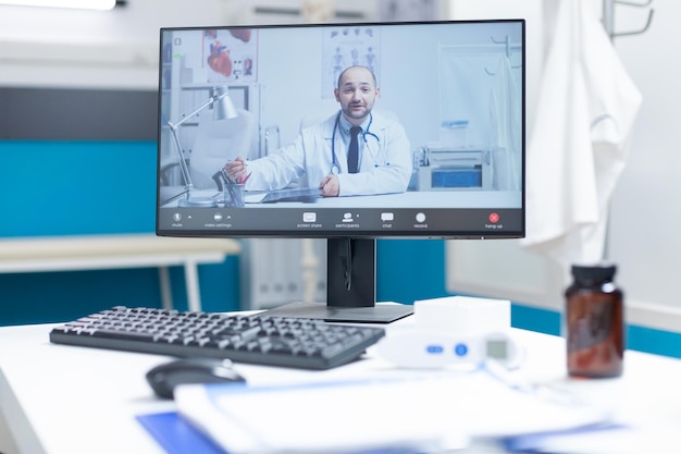 Computer met medische videocall-conferentievergadering op scherm op tafel in lege dokterspraktijk met niemand erin. Arts op afstand tijdens klinisch onderzoek. Bel voor ondersteuning in de gezondheidszorg