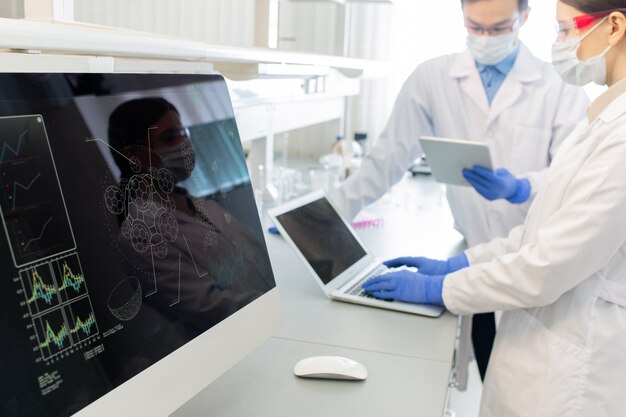 Foto schermo del computer con il modello della molecola sullo sfondo di due lavoratori di laboratorio in camici, maschere e guanti che studiano informazioni online