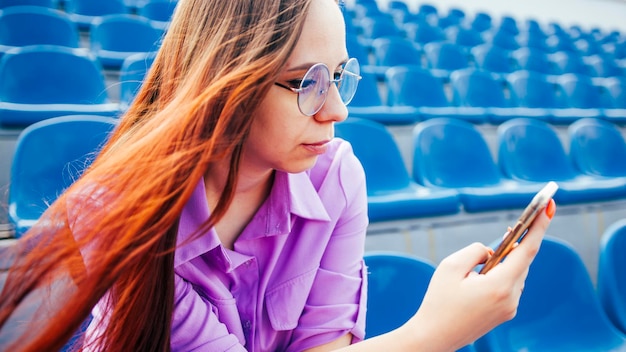 Concentrated adult female with long brown hair in blouse and eyeglasses sitting on seat of blue tribune and messaging on smartphone