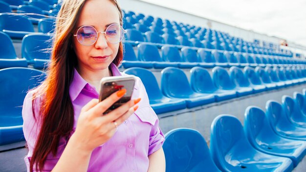 Concentrated adult female with long brown hair in blouse and eyeglasses sitting on seat of blue tribune and messaging on smartphone