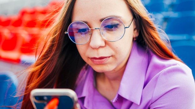 Concentrated adult female with long brown hair in blouse and eyeglasses sitting on seat of blue tribune and messaging on smartphone