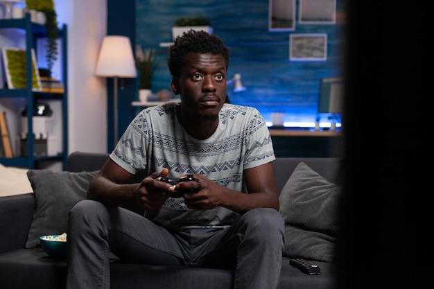Concentrated african american gamer playing online video games using gaming joystick during virtual championship. Competitive player enjoying free time sitting on sofa in living room