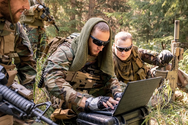 Programmatore dell'esercito concentrato in occhiali da sole che utilizza un computer militare mentre si entra in contatto con la base militare nella foresta