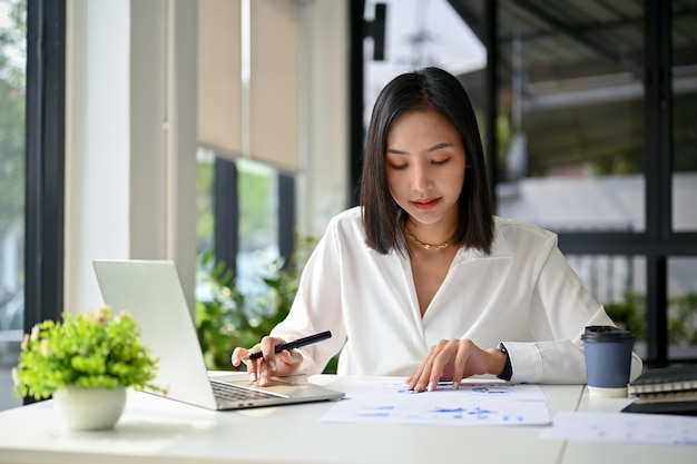 Concentrated Asian businesswoman analyzing financial data on reports working in her office