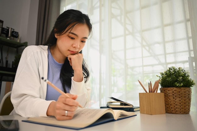 concentrated asian fe male student reading book preparing for exam or doing homework remotely at home