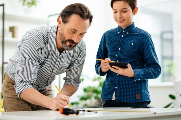 Concentrated bearded middle aged man drawing on the table and his smiling son standing near with modern tablet in his hands