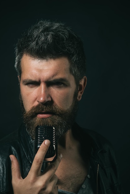 Concentrated bearded singer singing in microphone. talented\
handsome man in black leather jacket holds microphone and singing.\
rock star man with long beard. male singer with old fashioned\
microphone.