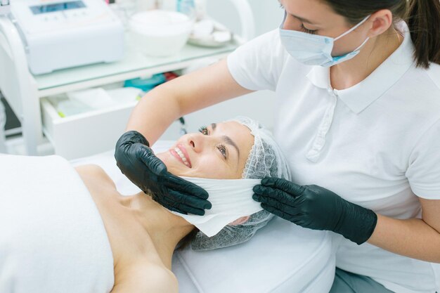 Concentrated beautician in a medical mask and rubber gloves touching the cheek of a contented client with a clean napkin