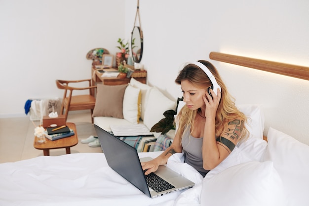 Сосредоточенная красивая молодая женщина-блогер в наушниках сидит в удобной кровати и редактирует видео на ноутбуке, выбирая хорошую музыку и звуковые эффекты