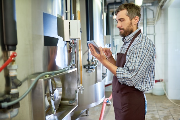 Concentrated beer engineer using tablet while examining equipment