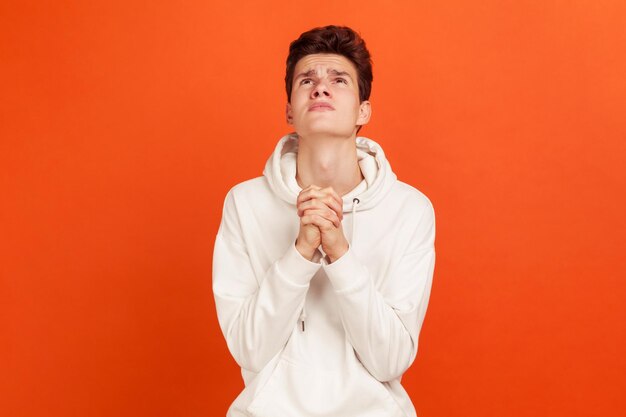 Photo concentrated believing teenager in casual sweatshirt crossing fingers of hands and rising eyes, praying god, asking for luck and blessing, faith. indoor studio shot isolated on orange background