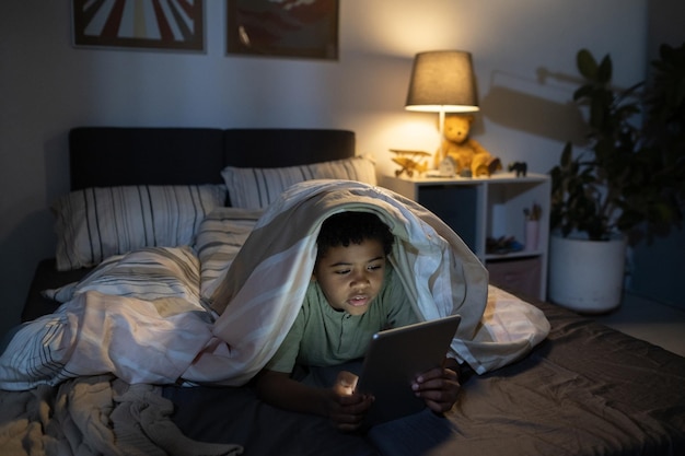 Concentrated black boy lying under duvet and using tablet in dark nursery room