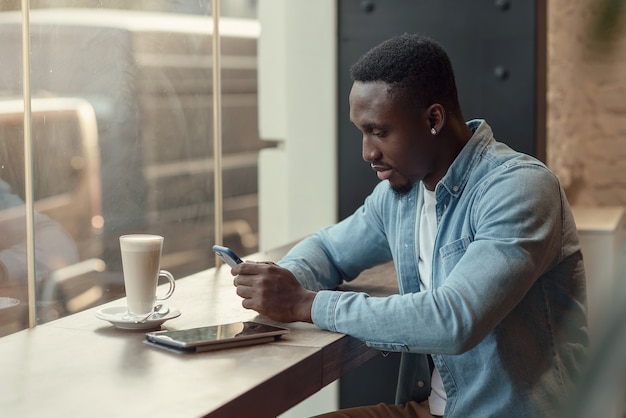 집중된 흑인 남성 기업가는 창문 가까이에 커피와 함께 카페에 앉아 스마트 폰을 사용합니다.
