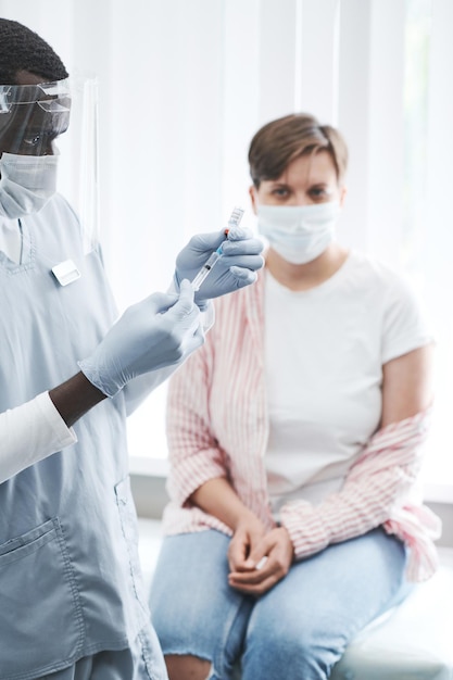 Концентрированная черная медсестра в защитном щитке для лица и перчатках готовит шприц перед вакцинацией от коронавируса