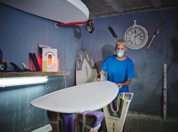 Concentrated blond haired male shaper in respirator mask examining shape of board placed on racks while creating handmade surfboards in workshop