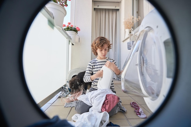 Concentrated boy with laundry and fabric softener near washing\
machine