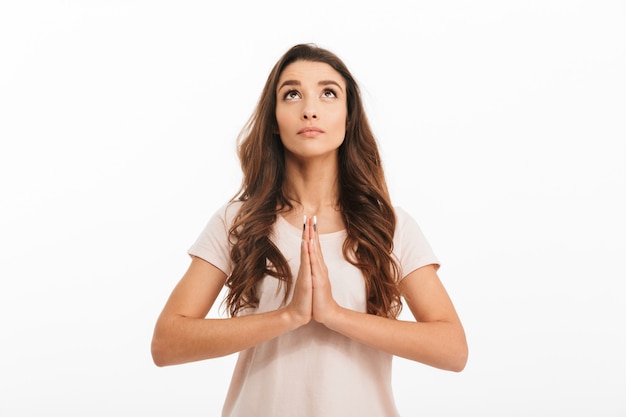 Concentrated brunette woman in t-shirt praying with pray gesture and looking up over white wall