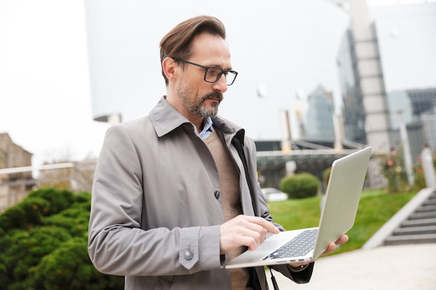 concentrated businessman in eyeglasses using laptop while standing at city street