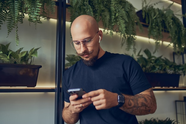 Concentrated businessman uses smartphone with headphones while stands in home office distance job