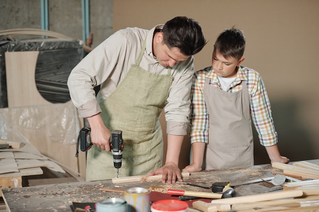 Concentrated carpenter in apron drilling wooden plank at desk while teenage son looking at its process in workshop