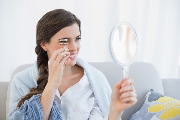 Concentrated casual brown haired woman in white pajamas using an eyelash curler