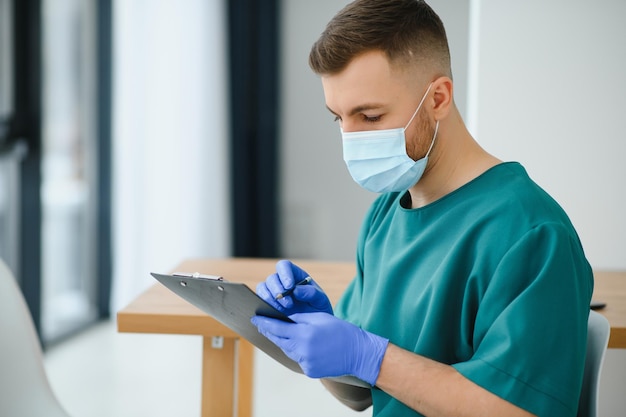 Concentrated caucasian doctor wearing protective mask with stethoscope holding clipboard and writing something