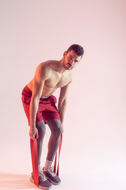 Photo concentrated caucasian sportsman doing exercise with resistance band. young handsome muscular bearded man with naked sportive torso. isolated on beige background. studio shoot. copy space