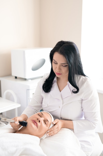 Cosmetologo concentrato in camice bianco utilizzando macchina per il viso mentre si esegue la procedura di rafforzamento della pelle per donna matura