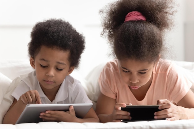 Concentrated cute little boy and girl laying on bed using\
gadgets reading studying playing games watching cartoons or movies\
scrolling social networks tech addicted children using devices