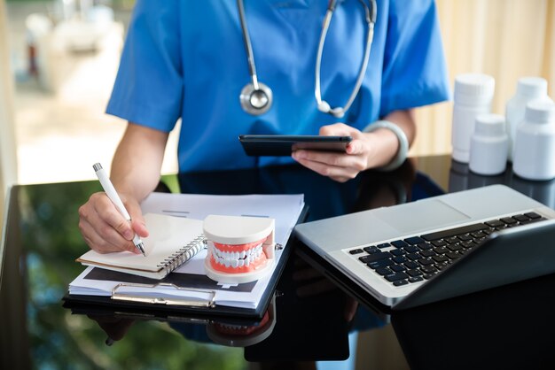 Concentrated dentist sitting at table with jaw samples tooth model and working with tablet and laptop in dental office professional dental clinic.