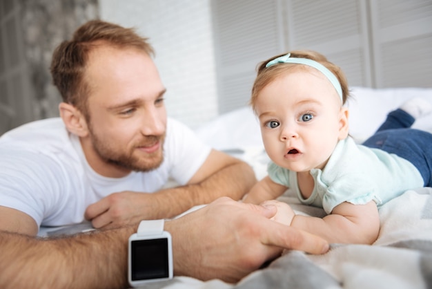 Concentrated on details around. Attentive charming positive baby girl lying on the bed with her father and looking on him while expressing interest and joy