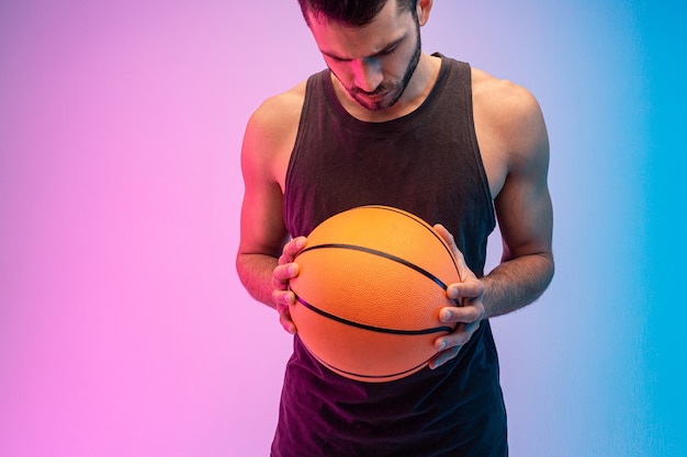 Concentrated european man hold basketball ball in hands. Young bearded guy wear tank top and looking down. Isolated on blue and pink background. Studio shoot. Copy space