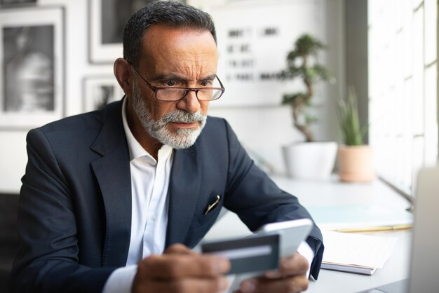 Concentrated european senior businessman in suit glasses use smartphone credit card at table in