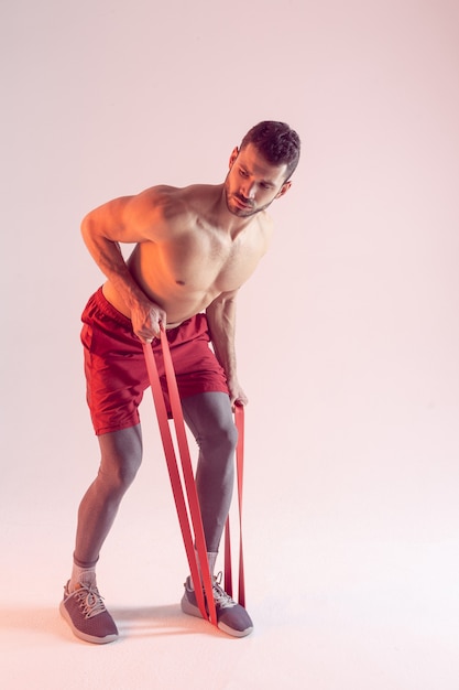 Концентрированный европейский спортсмен выполняет упражнения с лентой сопротивления. Молодой красивый мускулистый бородатый мужчина с обнаженным спортивным торсом. Изолированные на бежевом фоне. Студийная съемка. Копировать пространство