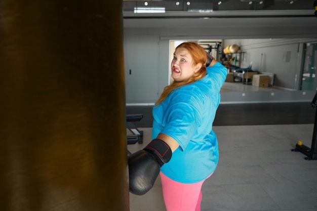 Concentrated fat woman using boxing gloves and bag doing punching exercises in gym. Sports motivation and healthy lifestyle