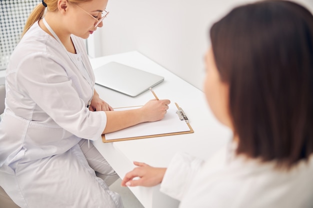 Concentrated female dermatologist in a lab coat writing with a pencil on a blank sheet of paper