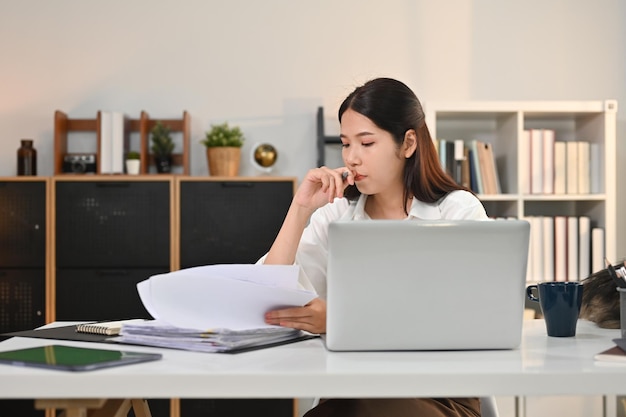 concentrated female siting in front of laptop computer at working desk and checking marketing reports