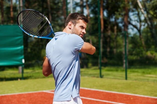 Concentrated on game. Handsome young man in polo shirt holding tennis racket and looking concentrated while standing on tennis court