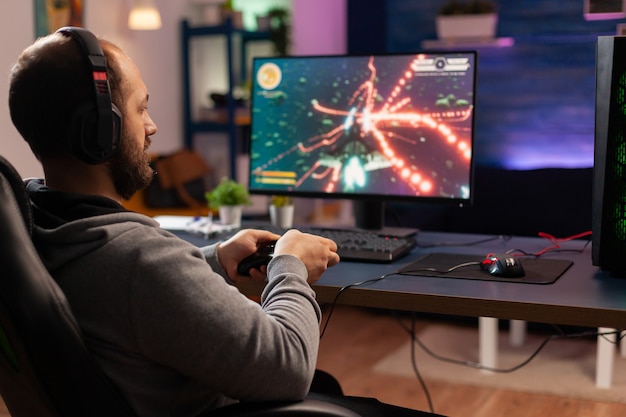 Concentrated gamer playing virtual game on powerful computer at\
home with professional headphones. digital gamer using joystick for\
shooter space gaming competition late at night in living room