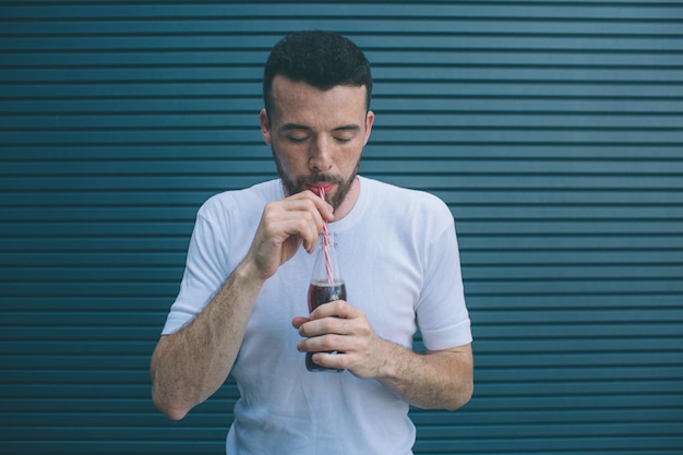 Concentrated guy is drinking coca-cola from bottle through straw. He is looking down. Isolated on striped 