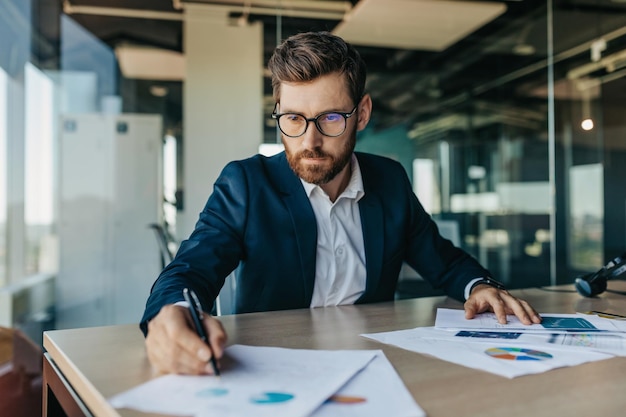 Foto uomo d'affari bello concentrato con gli occhiali che lavora con i documenti negli interni dell'ufficio per il controllo e la firma dei documenti