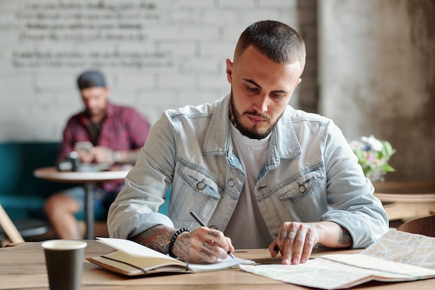 Ragazzo hipster concentrato con la barba seduto al tavolo in un caffè moderno e visualizzare la mappa cartacea mentre prende appunti sul viaggio